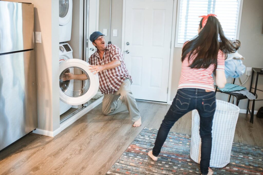 Man kneeling with back strain while using a front-load washing machine as woman brings laundry basket